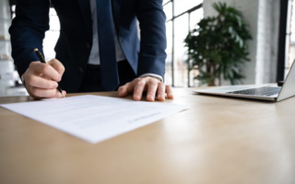 Close up young male ceo executive manager standing near table, doing company paperwork, putting signature on legal document, making corporate financial approval or signing business agreement.